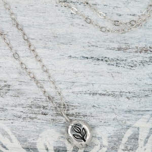 Puede incluir: Un collar de cadena de plata con un peque&ntilde;o colgante redondo con un dise&ntilde;o de hoja. El colgante es texturizado y tiene un aspecto r&uacute;stico.