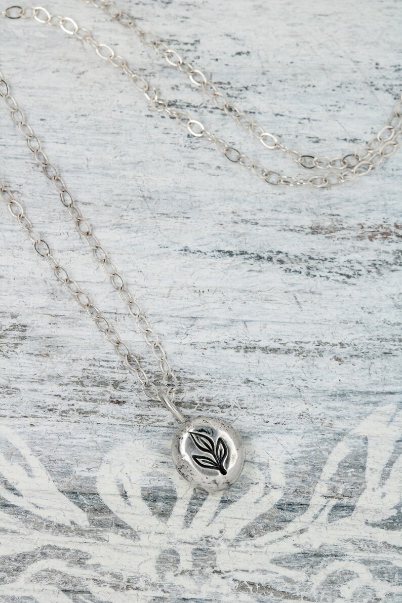 Puede incluir: Un collar de cadena de plata con un peque&ntilde;o colgante redondo con un dise&ntilde;o de hoja. El colgante es texturizado y tiene un aspecto r&uacute;stico.