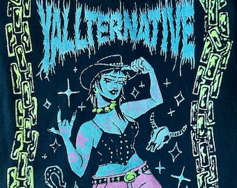May include: A black t-shirt with a green and blue graphic design. The graphic features a woman wearing a cowboy hat, a black corset, and pink pants. The text "Y ALTERNATIVE" is written in a stylized font above the woman.