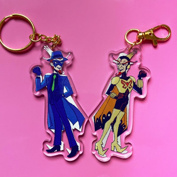 May include: Two colorful character keychains with gold chains. One keychain features a blue and green character with a mask and a hat. The other keychain features a yellow and red character with a butterfly design and a crown.