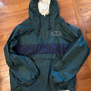 Può includere: Una giacca a vento con cappuccio verde scuro con una striscia orizzontale blu navy sul petto. La giacca ha una fodera bianca nel cappuccio e le lettere greche "AXA" sono ricamate sul davanti. La giacca ha un cordoncino sul cappuccio e sull'orlo.