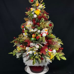 May include: A small artificial Christmas tree decorated with red, white, and gold ornaments, including a few rubber duckies. The tree is in a red pot with a white fur trim and is sitting on a white pedestal.
