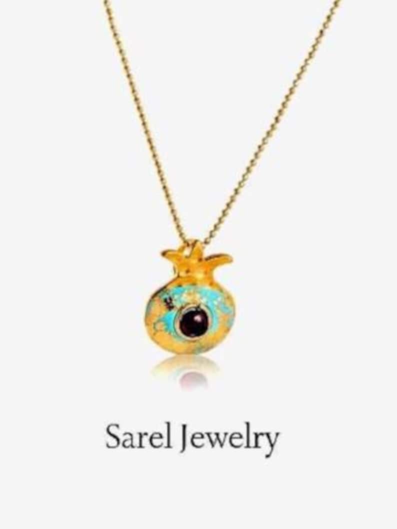 Peut inclure: Un collier dor&eacute; avec un pendentif en forme de grenade. Le pendentif est orn&eacute; d'&eacute;mail turquoise et d'une pierre rouge fonc&eacute; au centre. Le texte "Sarel Jewelry" est visible en dessous.