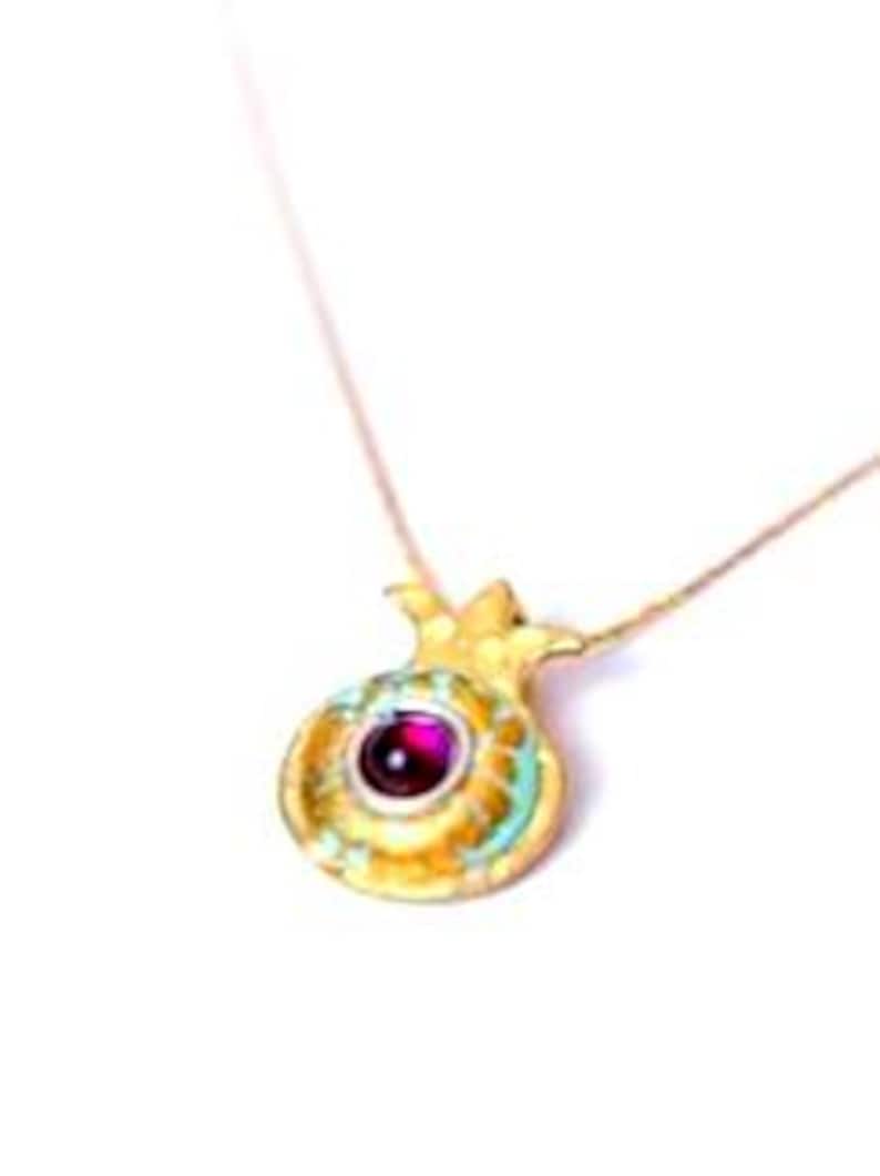 Peut inclure: Un collier pendentif dor&eacute; repr&eacute;sentant une grenade. Le pendentif est orn&eacute; d'une pierre pr&eacute;cieuse rouge ronde au centre et de d&eacute;tails turquoise. La cha&icirc;ne du collier est fine et dor&eacute;e.