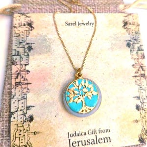 May include: A gold-tone chain necklace with a circular pendant featuring a gold tree silhouette set against a bright turquoise background. The pendant is displayed on a card labeled Sarel Jewelry and Judaica Gift from Jerusalem.