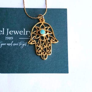 Peut inclure: Un collier avec un pendentif main de Hamsa doré présentant un design en filigrane complexe et une petite pierre ronde de couleur bleu turquoise au centre. Le pendentif est suspendu à une fine chaîne dorée sur une carte vert foncé.