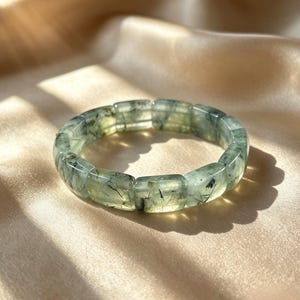 May include: A light green prehnite bracelet with rectangular beads. The bracelet has dark inclusions and is displayed on a gold satin surface. The gemstone bracelet is a piece of jewelry.