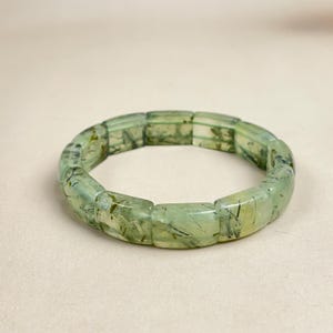 May include: A green prehnite bracelet. The bracelet is made of rectangular, translucent beads with dark green and black inclusions. The beads are strung together to form a circular shape. The bracelet is displayed on a neutral background.