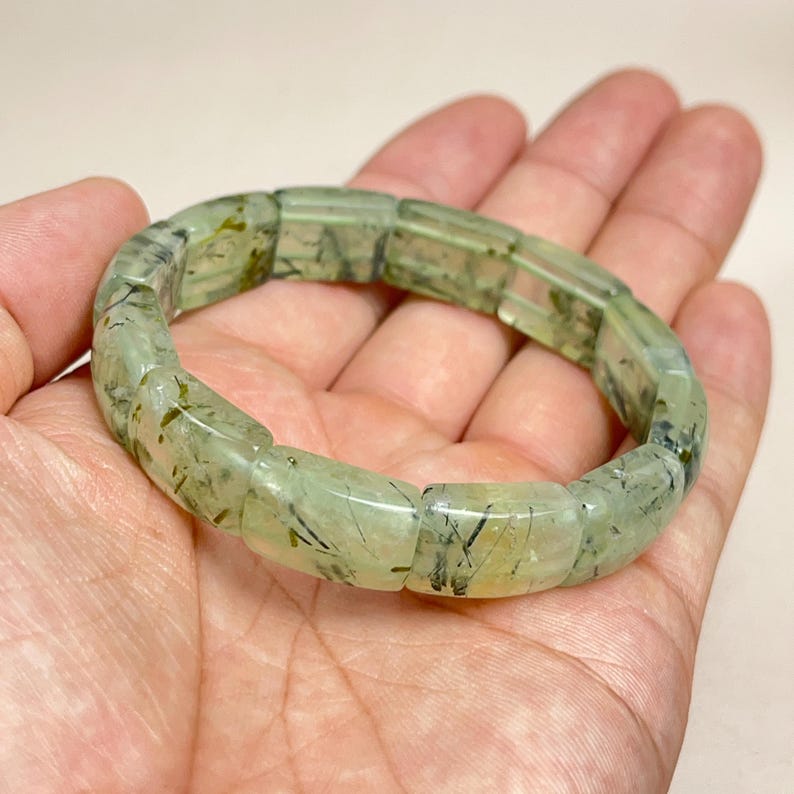 May include: A light green prehnite bracelet. The bracelet is composed of rectangular, translucent beads with dark green inclusions. The bracelet is held in a hand, showcasing the natural stone's color and pattern.