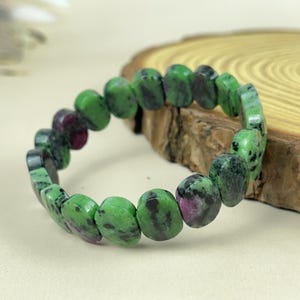 Op de afbeelding: Een armband met ovale stenen in groen en kastanjebruin. De stenen zijn aan elkaar geregen tot een complete cirkel. De armband wordt gepresenteerd op een houten oppervlak.