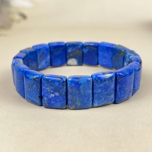 May include: A vibrant blue lapis lazuli bracelet. The bracelet is composed of rectangular, polished stones with gold flecks. The stones are strung together to form a flexible band. The jewelry is displayed on a neutral background.