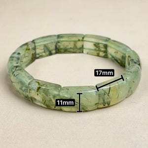 May include: A light green prehnite bracelet. The bracelet is made of rectangular beads, each measuring 11mm x 17mm. The beads have a translucent quality with darker green inclusions. The bracelet is displayed on a neutral background.