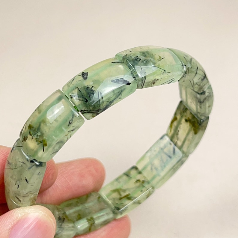 May include: A light green prehnite bracelet. The bracelet is composed of rectangular, translucent beads with dark green and black inclusions. The beads are strung together to form a flexible band. The bracelet is held in a hand.