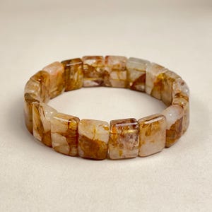 May include: A square-beaded bracelet featuring translucent, amber-colored stones with brown inclusions. The bracelet is made of polished, rectangular beads strung together, showcasing a natural, earthy aesthetic. The bracelet is designed to be worn around the wrist.