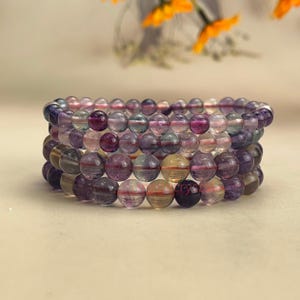 Op de afbeelding: Een stapel van vier armbanden gemaakt met regenboogfluoriet kralen. De kralen zijn rond en hebben een verscheidenheid aan kleuren, waaronder paars, roze, groen en geel.