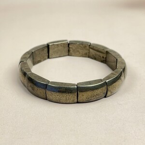 May include: A silver-toned bracelet with rectangular links. The bracelet has a slightly tarnished appearance, suggesting an aged or vintage aesthetic. The links are connected to form a flexible band, suitable for wrist wear.