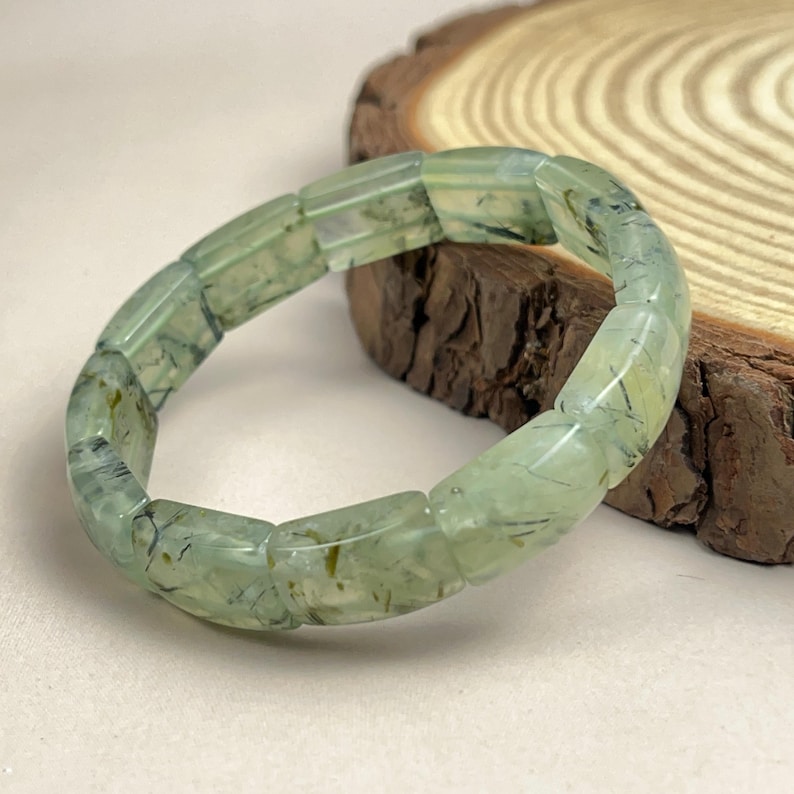 May include: A light green prehnite bracelet. The bracelet is made of rectangular, translucent beads with dark green inclusions. The beads are strung together to form a circular shape. The bracelet is displayed on a light-colored surface.