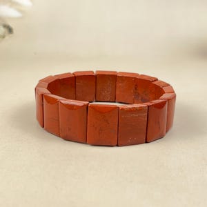 以下が含まれることがあります： 赤いジャスパーのブレスレット。ブレスレットは、長方形で研磨された石でできています。石は深紅の色で、微妙な色のバリエーションがあります。ブレスレットは手首に着用するように設計されています。