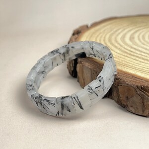 May include: A clear quartz bracelet with black tourmaline inclusions. The bracelet is composed of rectangular, faceted beads. The black inclusions create a striking contrast against the translucent quartz. The bracelet is displayed on a wooden surface.
