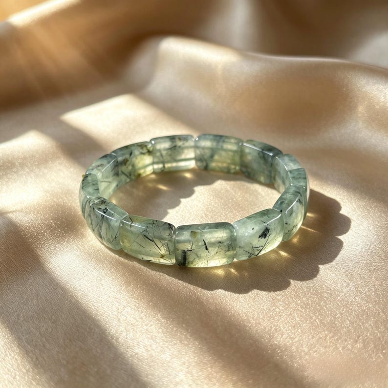 May include: A light green prehnite bracelet with rectangular beads. The bracelet has dark inclusions and is displayed on a gold satin surface. The gemstone bracelet is a piece of jewelry.