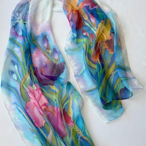 May include: A long, rectangular scarf featuring a hand-painted floral design of purple, pink, and orange iris flowers with green stems and leaves. The background is a soft, blended gradient of light blue and white, creating a fluid, airy appearance.