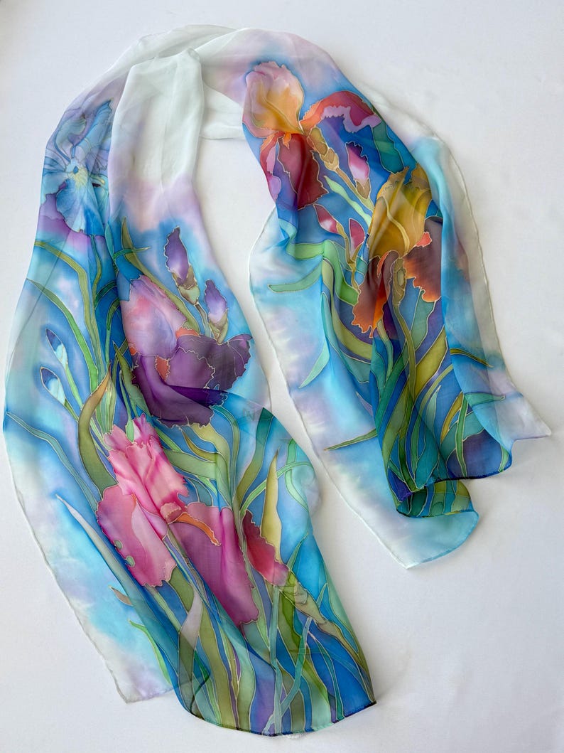 May include: A long, rectangular scarf featuring a hand-painted floral design of purple, pink, and orange iris flowers with green stems and leaves. The background is a soft, blended gradient of light blue and white, creating a fluid, airy appearance.