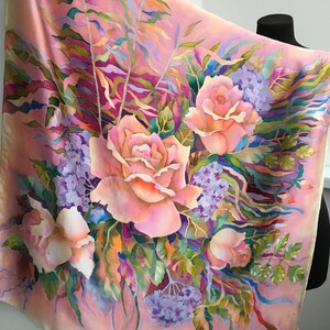 May include: A square silk scarf featuring a hand-painted floral design on a soft peach background. The pattern displays large, light pink roses and clusters of small purple flowers, accented by flowing green and teal foliage in a watercolor style.
