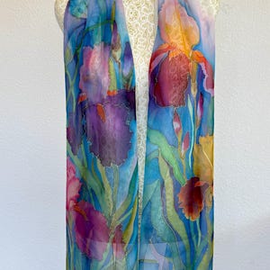 May include: A long, rectangular scarf featuring a hand-painted floral design of purple, blue, and orange irises with green stems and leaves. The fabric is semi-sheer with a smooth texture, displaying vibrant watercolor-style botanical illustrations.