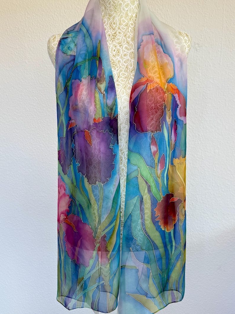 May include: A long, rectangular scarf featuring a hand-painted floral design of purple, blue, and orange irises with green stems and leaves. The fabric is semi-sheer with a smooth texture, displaying vibrant watercolor-style botanical illustrations.