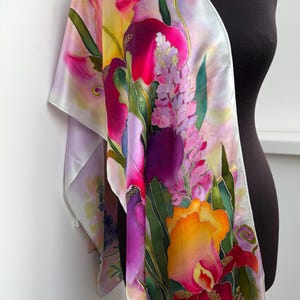 K&ouml;nnte beinhalten: Ein Seidenschal mit einem lebendigen, handbemalten Blumendesign mit gro&szlig;en Schwertlilien und Lupinen in Magenta-, Violett-, Gelb- und Oranget&ouml;nen. Der Hintergrund ist in zartem Lavendel und Wei&szlig; gehalten, mit gr&uuml;nen Blattakzenten auf dem Stoff.