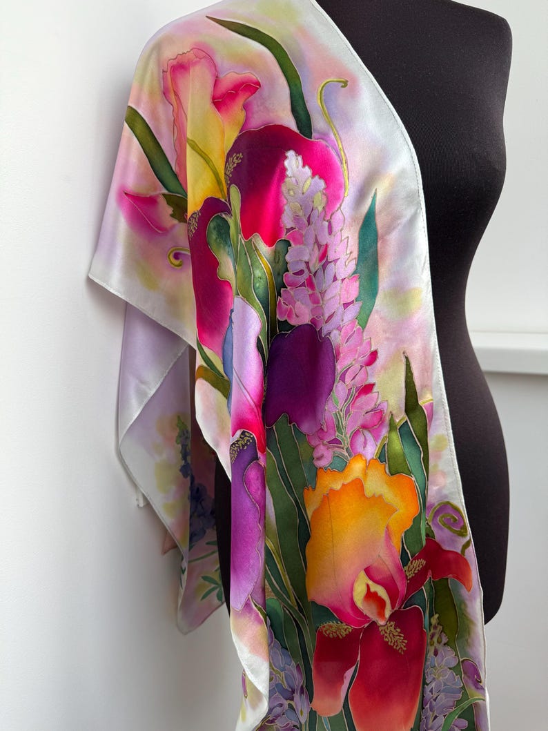 K&ouml;nnte beinhalten: Ein Seidenschal mit einem lebendigen, handbemalten Blumendesign mit gro&szlig;en Schwertlilien und Lupinen in Magenta-, Violett-, Gelb- und Oranget&ouml;nen. Der Hintergrund ist in zartem Lavendel und Wei&szlig; gehalten, mit gr&uuml;nen Blattakzenten auf dem Stoff.