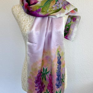 K&ouml;nnte beinhalten: Ein rechteckiger Seidenschal mit einem handgemalten Blumenmuster aus hohen violetten und rosafarbenen Lupinen vor einem zarten Pastellhintergrund in Hellrosa, Gelb und Gr&uuml;n. Der Schal hat eine glatte, gl&auml;nzende Oberfl&auml;che und fallende Kanten.
