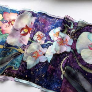Puede incluir: Una bufanda de seda con un diseño pintado a mano de orquídeas blancas sobre un fondo morado y azul con detalles dorados.