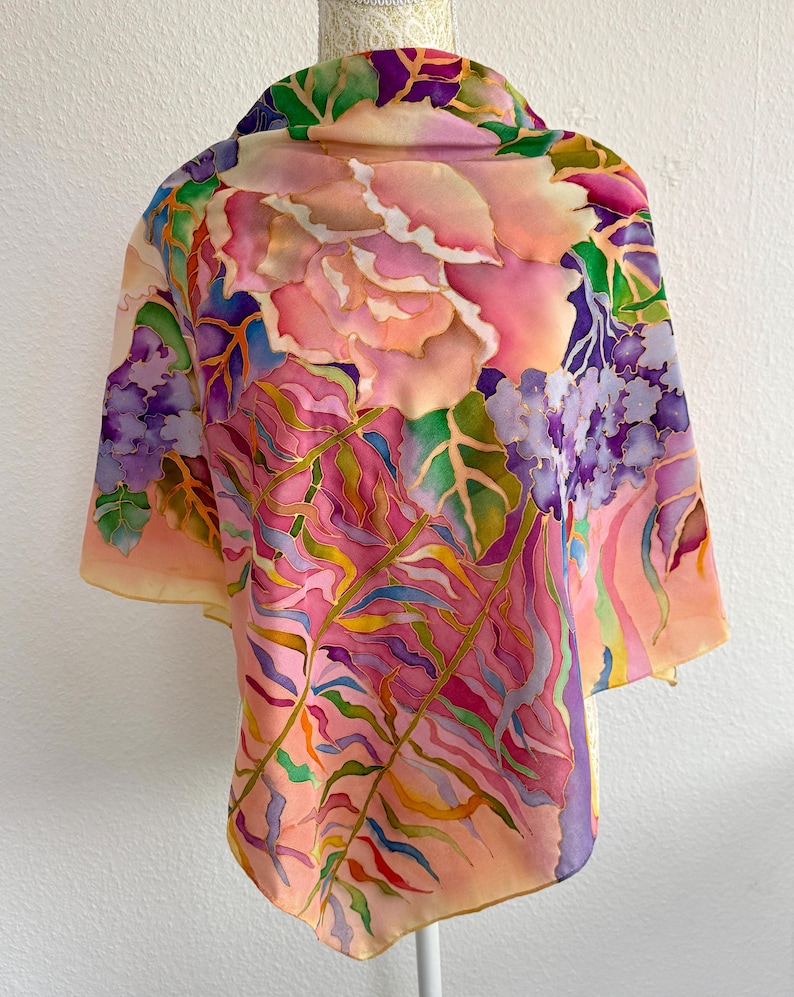 May include: A lightweight scarf featuring a hand-painted floral design with large pink roses, purple hydrangea clusters, and long green fern fronds. The fabric displays a vibrant color palette of pink, purple, green, and orange hues with defined outlines.