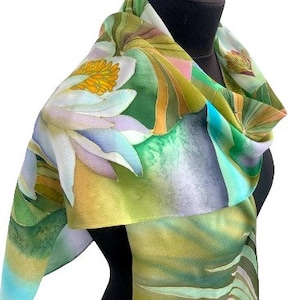 Puede incluir: Una bufanda de seda con un dise&ntilde;o floral verde, azul y blanco. La bufanda presenta una gran flor blanca con un centro amarillo y hojas verdes. La bufanda est&aacute; doblada y drapeada sobre un maniqu&iacute;.