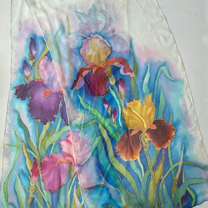 May include: A rectangular silk scarf featuring a hand-painted floral design of multiple iris flowers in shades of deep purple, magenta, and golden yellow. The flowers are set against a vibrant blue background with green foliage and a white border.