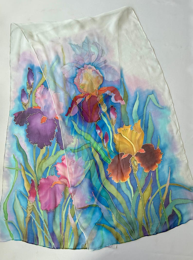 May include: A rectangular silk scarf featuring a hand-painted floral design of multiple iris flowers in shades of deep purple, magenta, and golden yellow. The flowers are set against a vibrant blue background with green foliage and a white border.