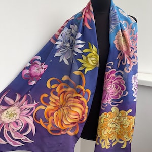 May include: A silk scarf with a purple, blue, and yellow gradient background. The scarf is decorated with hand-painted chrysanthemum flowers in shades of pink, orange, and yellow.