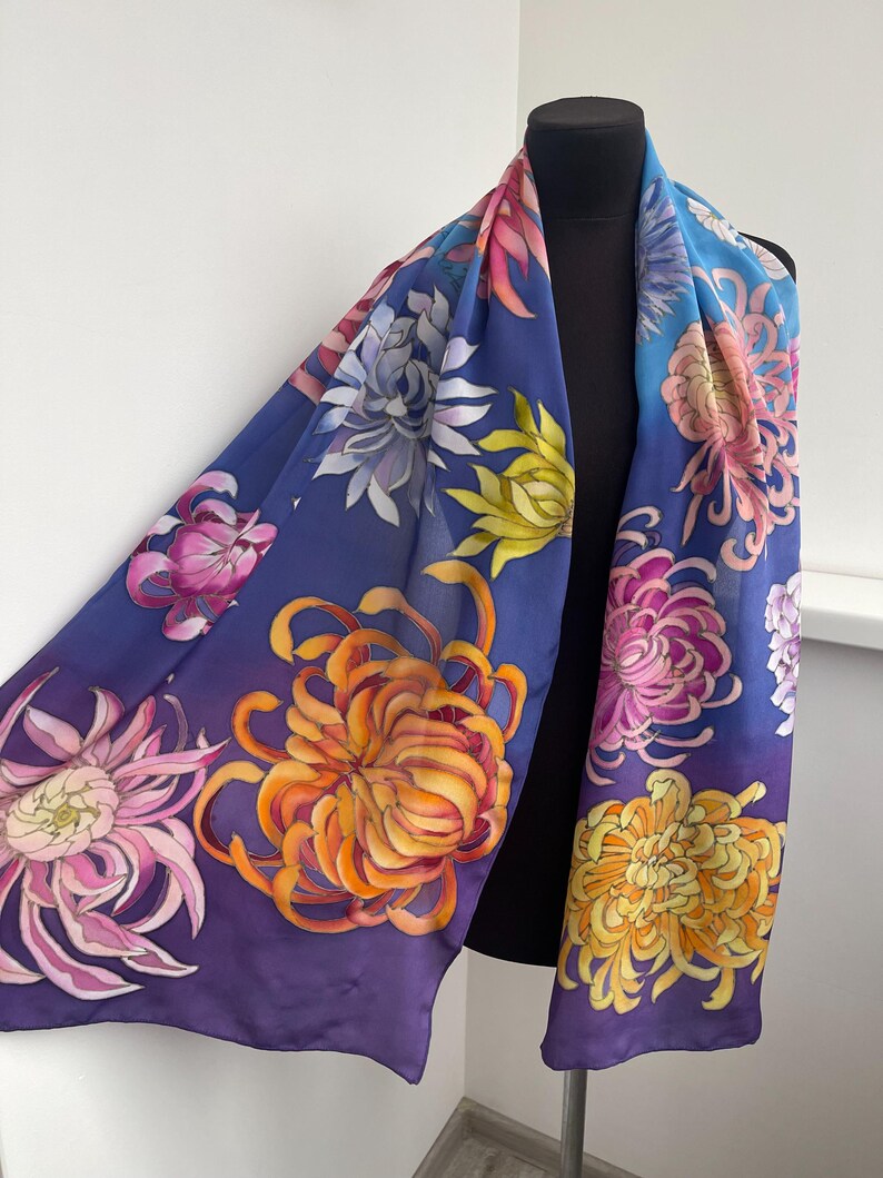 May include: A silk scarf with a purple, blue, and yellow gradient background. The scarf is decorated with hand-painted chrysanthemum flowers in shades of pink, orange, and yellow.
