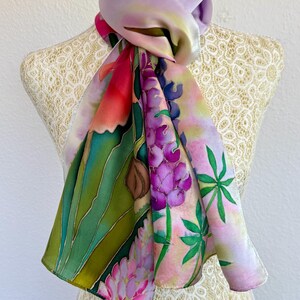 K&ouml;nnte beinhalten: Ein handbemaltes Seidentuch mit einem lebendigen botanischen Design aus violetten Lupinenbl&uuml;ten, gr&uuml;nem Blattwerk und zarten rosa Akzenten. Das Tuch zeigt eine glatte, gl&auml;nzende Textur mit einem lavendelfarbenen Farbverlauf am Halsbereich.