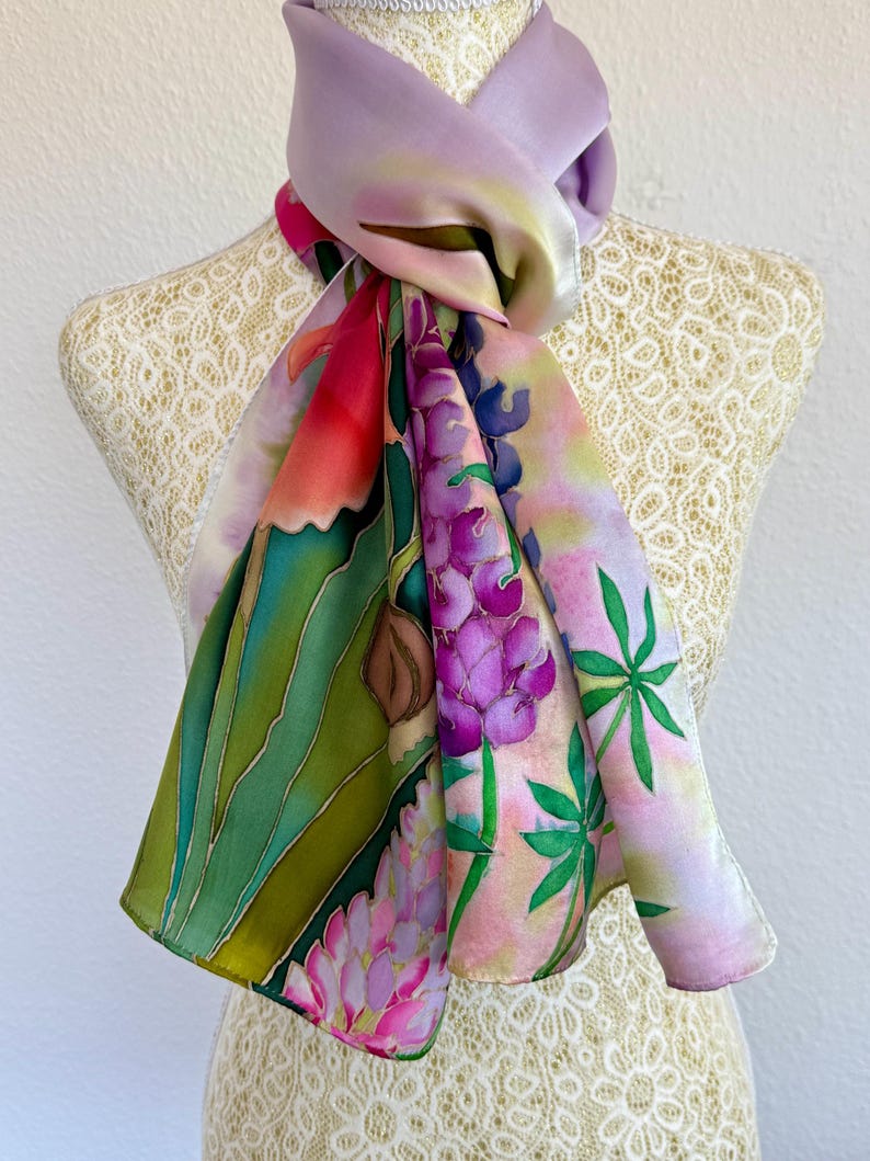 K&ouml;nnte beinhalten: Ein handbemaltes Seidentuch mit einem lebendigen botanischen Design aus violetten Lupinenbl&uuml;ten, gr&uuml;nem Blattwerk und zarten rosa Akzenten. Das Tuch zeigt eine glatte, gl&auml;nzende Textur mit einem lavendelfarbenen Farbverlauf am Halsbereich.
