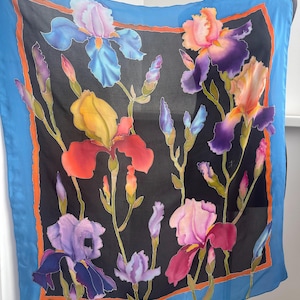 May include: A silk scarf with a black background and a colorful floral design featuring irises. The irises are painted in shades of purple, blue, yellow, orange, and red. The scarf has a blue border with an orange inner border.