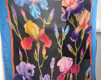 May include: A silk scarf with a black background and a colorful floral design featuring irises. The irises are painted in shades of purple, blue, yellow, orange, and red. The scarf has a blue border with an orange inner border.