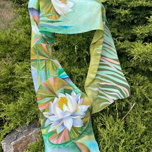 Puede incluir: Una bufanda de seda verde y blanca con un dise&ntilde;o floral. La bufanda presenta una gran flor blanca con un centro amarillo y hojas verdes. El fondo es de color verde claro con una franja blanca y rosa.