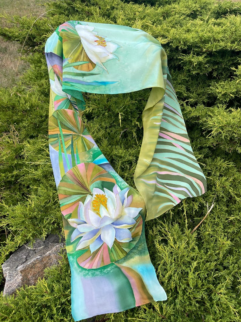 Puede incluir: Una bufanda de seda verde y blanca con un dise&ntilde;o floral. La bufanda presenta una gran flor blanca con un centro amarillo y hojas verdes. El fondo es de color verde claro con una franja blanca y rosa.