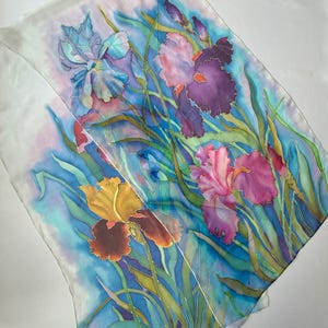 May include: A rectangular silk scarf featuring a hand-painted floral design of irises in shades of deep purple, magenta, and golden yellow. The flowers are surrounded by long green leaves against a vibrant blue and white watercolor-style background.