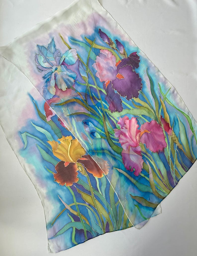 May include: A rectangular silk scarf featuring a hand-painted floral design of irises in shades of deep purple, magenta, and golden yellow. The flowers are surrounded by long green leaves against a vibrant blue and white watercolor-style background.