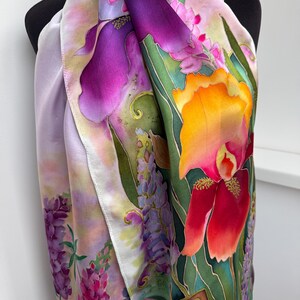 K&ouml;nnte beinhalten: Ein Seidenschal mit einem lebendigen Blumenmuster, das gro&szlig;e violette und orangefarbene Irisbl&uuml;ten, Gruppen kleinerer rosa und violetter lupinenartiger Bl&uuml;ten sowie gr&uuml;nes Blattwerk auf einem lavendel- und rosafarbenen Hintergrund zeigt.