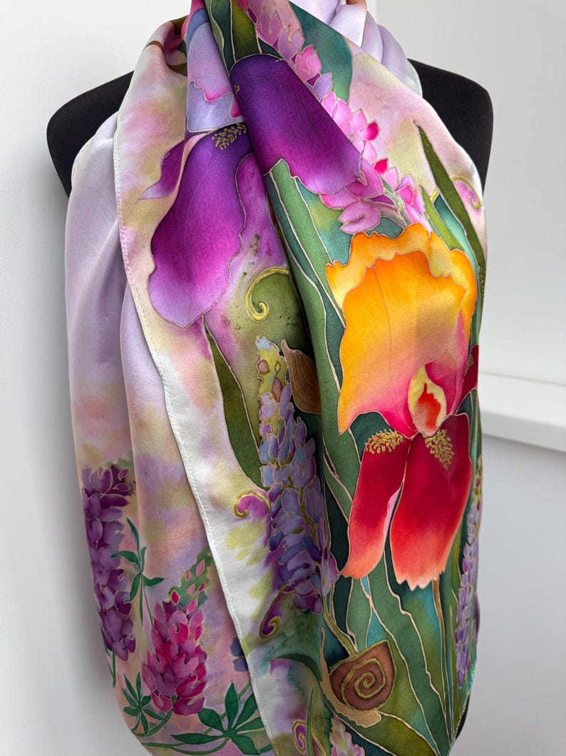 K&ouml;nnte beinhalten: Ein Seidenschal mit einem lebendigen Blumenmuster, das gro&szlig;e violette und orangefarbene Irisbl&uuml;ten, Gruppen kleinerer rosa und violetter lupinenartiger Bl&uuml;ten sowie gr&uuml;nes Blattwerk auf einem lavendel- und rosafarbenen Hintergrund zeigt.