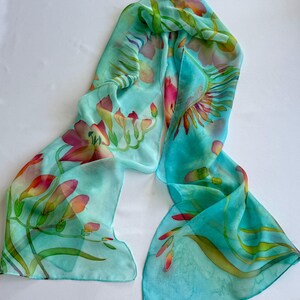 Pode incluir: Um len&ccedil;o retangular de seda com um fundo turquesa vibrante e motivos florais pintados &agrave; m&atilde;o em tons de rosa, laranja e amarelo. O design inclui caules e folhas verdes delicadas sobre um tecido leve e transl&uacute;cido.